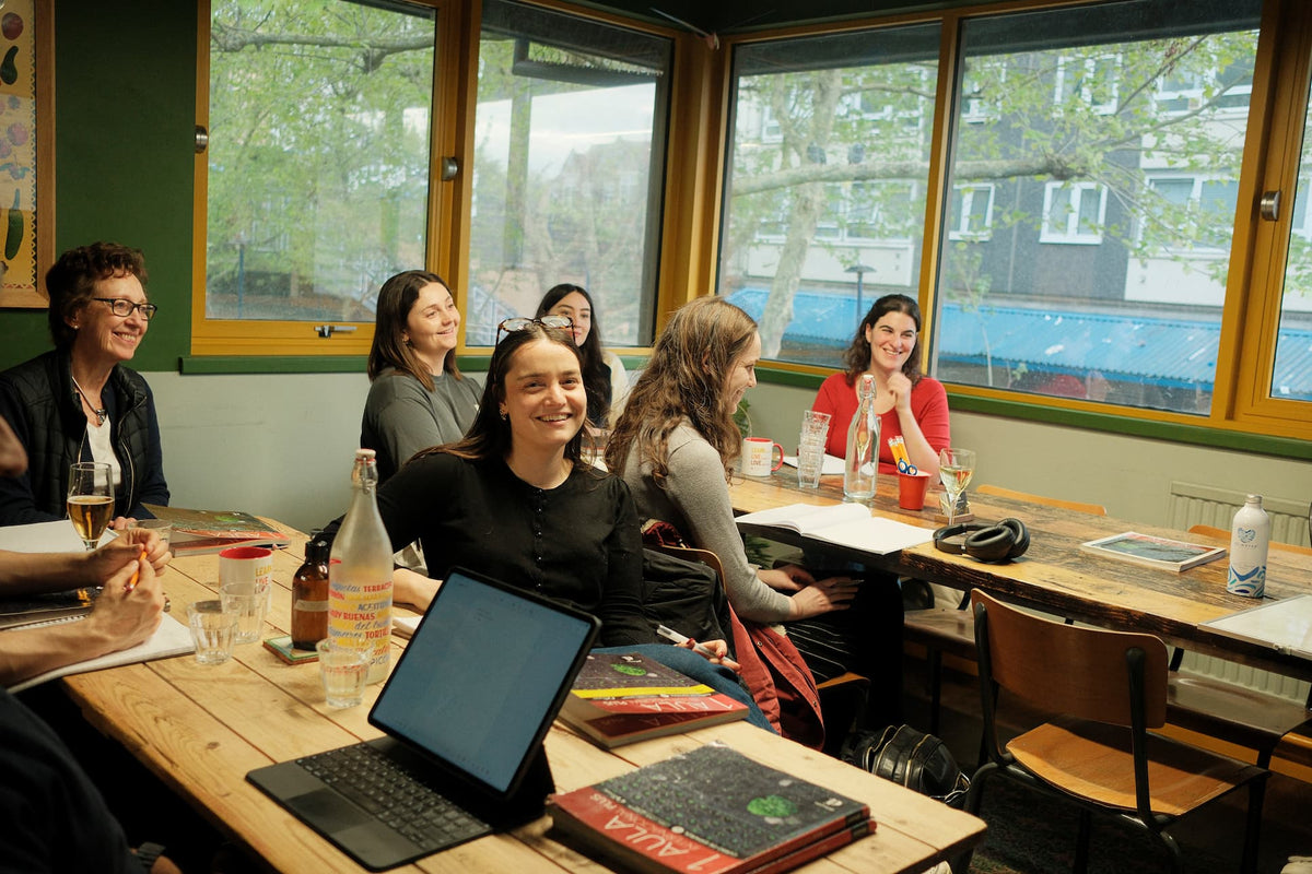 A group of students sit in a brightly decorated room, at rustic wooden tables, smiling and enjoying their lesson.