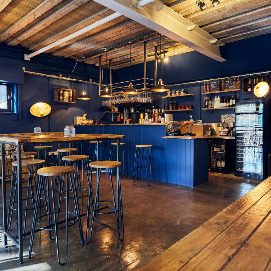 A dark blue bar and lounge interior with wooden ceiling beams, hanging pendant lights, bar stools, a counter, shelving, and a beverage refrigerator.