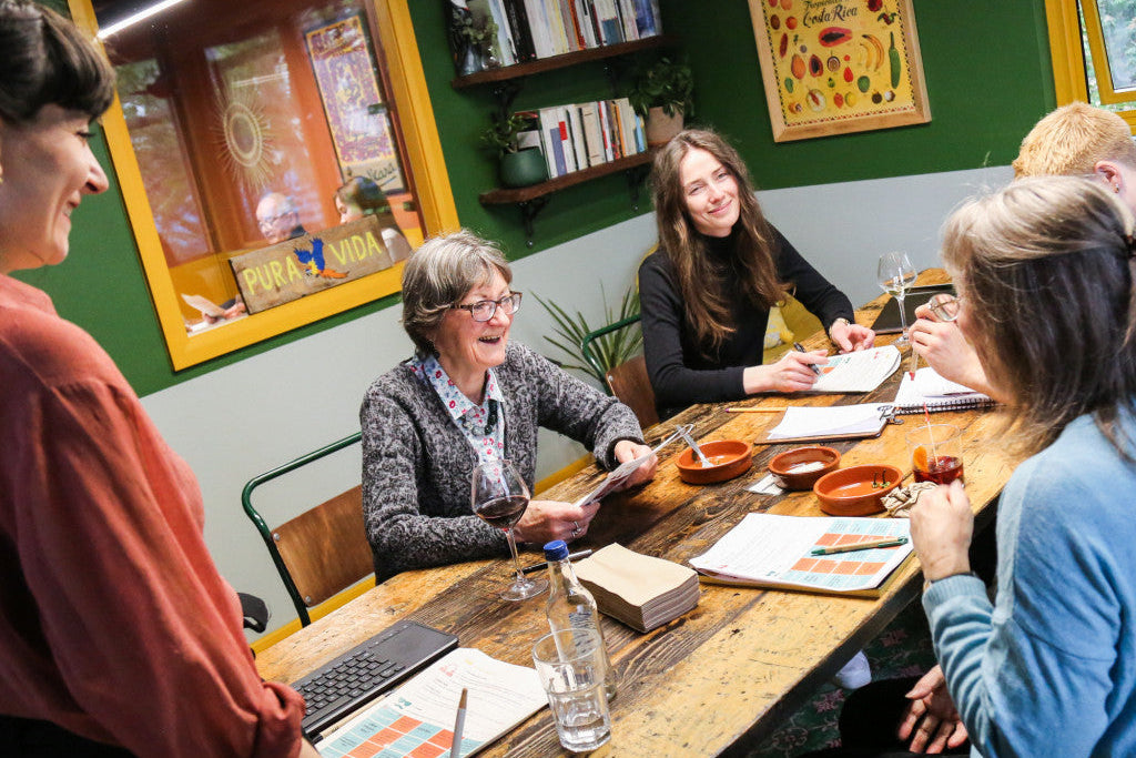 A group of five women gathered around a rustic wooden table in a cozy room with green walls, sharing a meal and engaging in conversation. The table is set with books, notebooks, a laptop, and small bowls of food.
