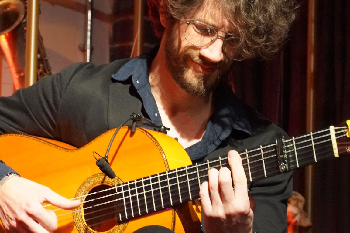 A man with glasses and a beard sits on a wooden stool, playing a classical acoustic guitar with a black shirt and black trousers in a warmly lit indoor setting.
