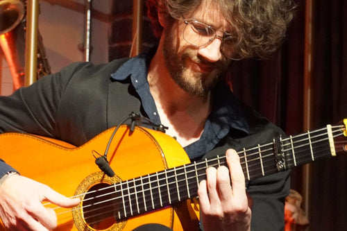 A man with glasses and a beard sits on a wooden stool, playing a classical acoustic guitar with a black shirt and black trousers in a warmly lit indoor setting.