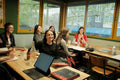 A group of students sit in a brightly decorated room, at rustic wooden tables, smiling and enjoying their lesson.
