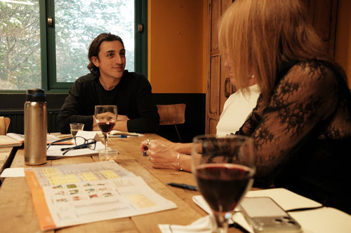 Two students sit looking at each other eagerly, at a rustic wooden table with textbooks and glasses of wine on the table.