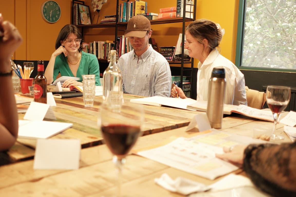 A group of people seated around a wooden table in a warm indoor setting, with books on shelves in the background. The scene includes wine glasses, bottles, notebooks, and a metal water bottle.