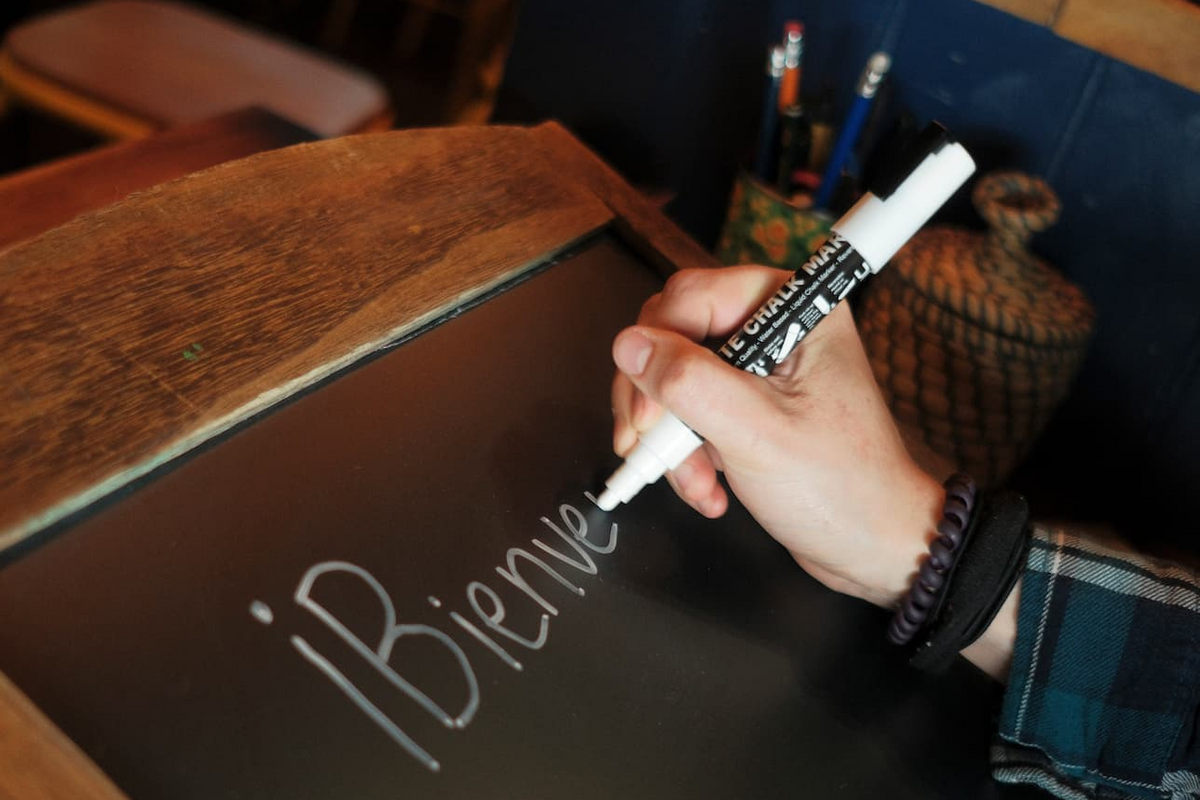 A hand holding a white marker writes on a blackboard, with the word 'Bienvenidos' partially visible. 