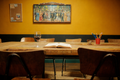 A rustic wooden dining table in a cozy room with a mustard yellow wall, featuring an open book, a glass of red wine, and chairs. A large framed painting hangs on the wall above the table.
