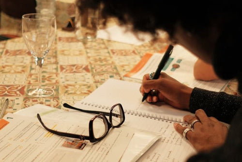 A close-up lifestyle shot of a person writing in a notebook at a table, with black-rimmed eyeglasses resting on papers in the foreground and a wine glass in the background.