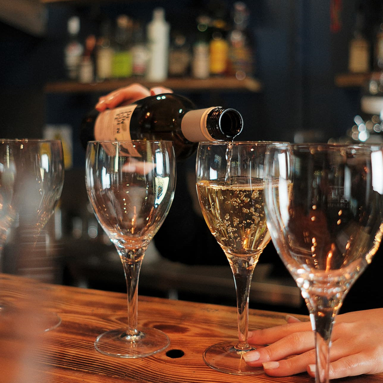 A hand pours white wine from a dark bottle into a stemmed glass on a wooden bar counter, with other empty glasses and a blurred bar background.