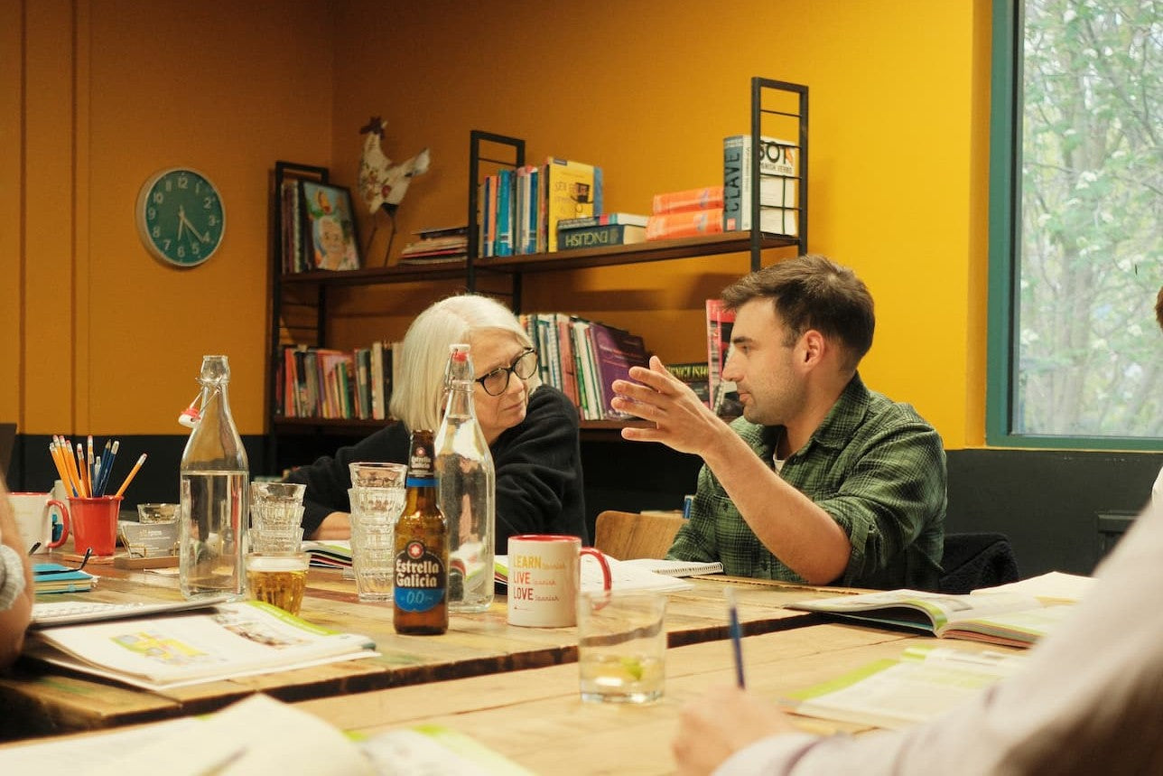 A man in a green shirt gestures while speaking to a woman with white hair and glasses seated at a wooden table in a warm, yellow-walled room with bookshelves in the background.