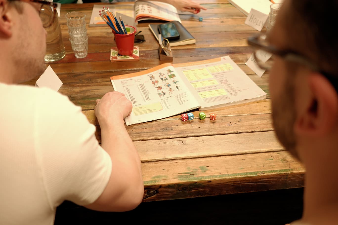 Two people seated at a rustic wooden table during a game session, looking at printed game instructions with colorful dice on the table.