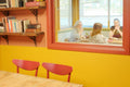 A warm interior scene with a yellow and orange wall, wooden bookshelves, two red chairs in the foreground, and a large window looking out onto three women seated at a table.