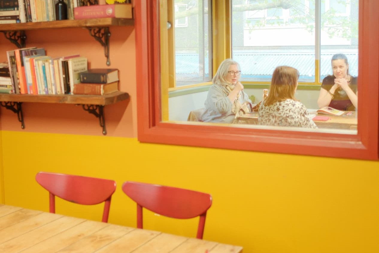 A warm interior scene with a yellow and orange wall, wooden bookshelves, two red chairs in the foreground, and a large window looking out onto three women seated at a table.
