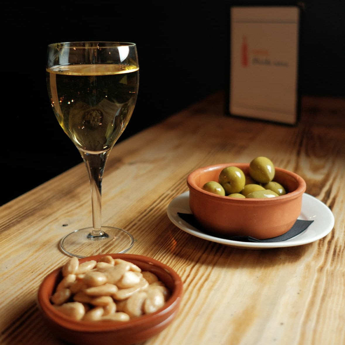 A glass of white wine on a wooden table alongside a small terracotta bowl of green olives on a white saucer and another small terracotta bowl of white beans, with a dark restaurant background.