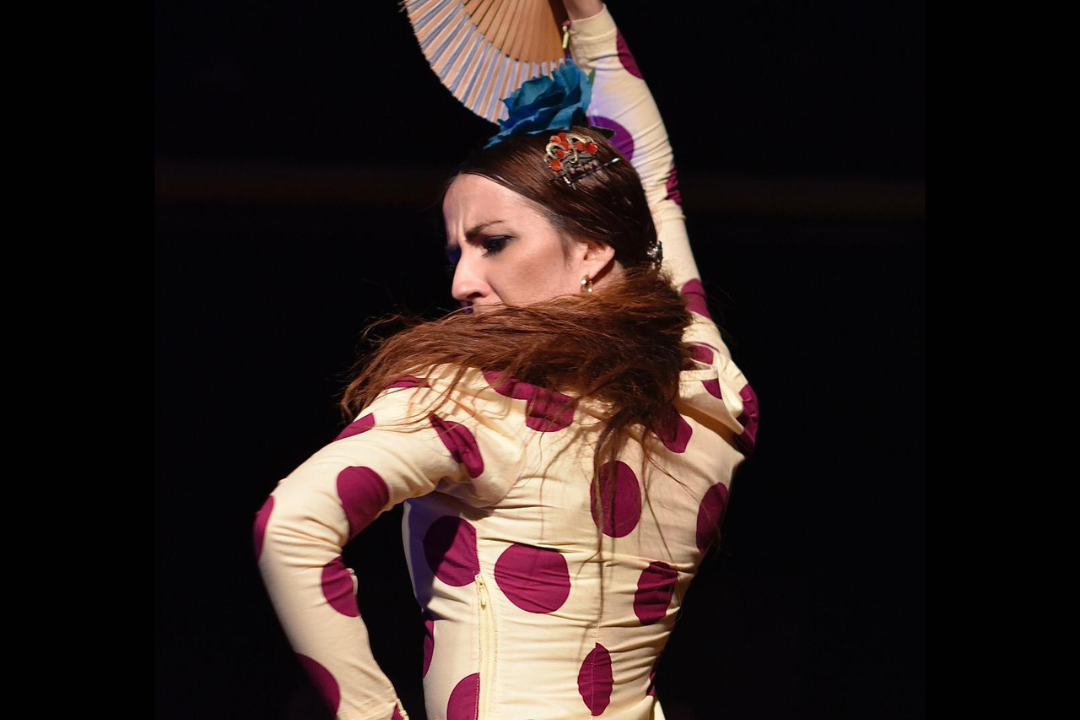 A woman in a long-sleeve cream dress with large dark magenta polka dots poses dynamically with a folding fan against a black background.