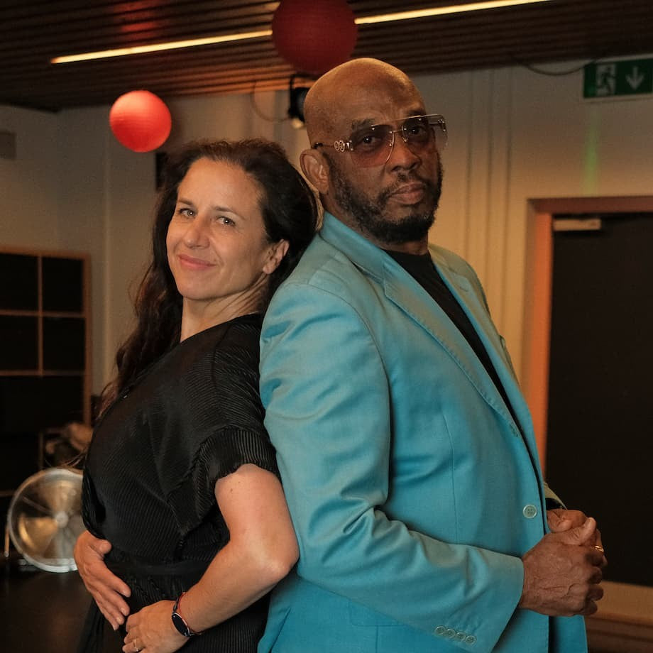 A man in a bright turquoise blazer and sunglasses standing arm-in-arm with a woman in a black dress in an indoor event space with red balloons.
