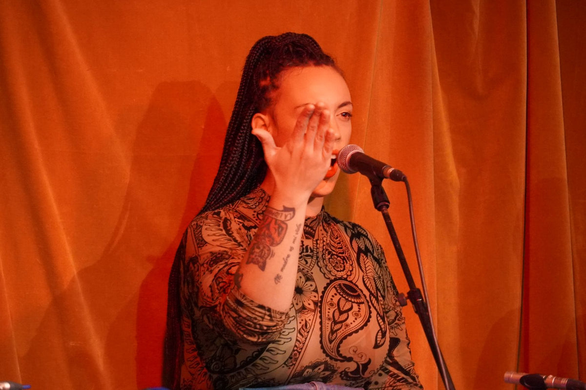A person with long dark braids stands in front of a microphone wearing a patterned black top, set against a draped orange curtain background.