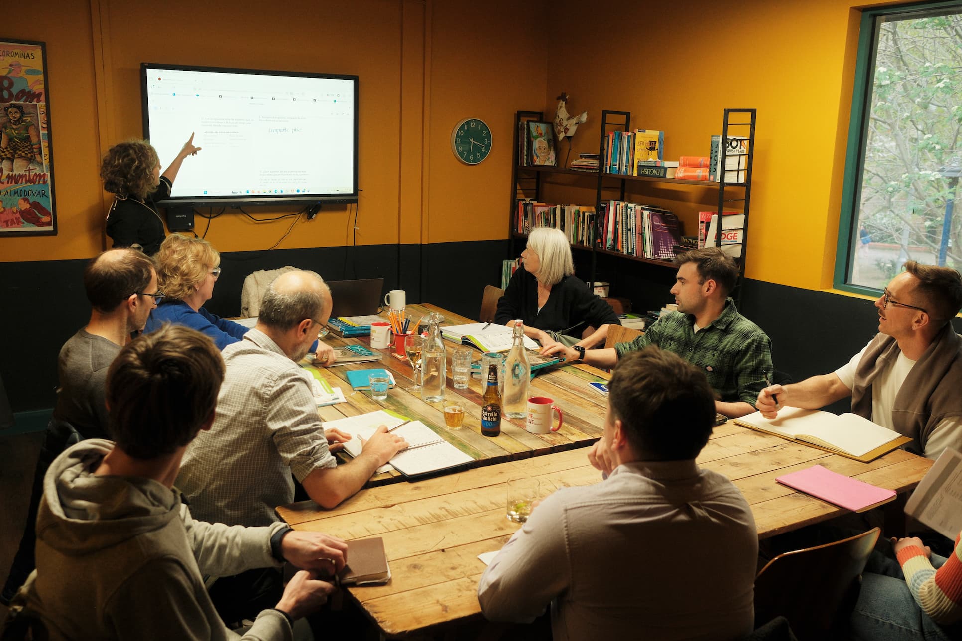 A group of people seated around a wooden table in a meeting room with orange walls, gathered around a large screen being pointed at by a presenter.