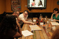 A group of people seated around a rustic wooden table in a dimly lit indoor space, looking at printed menus or papers. A television on the wall displays a video, and the room has warm orange-brown walls with posters and dark paneling.