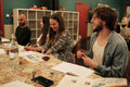 A group of four young adults sitting around a table in a classroom or workshop setting, smiling and laughing while reviewing printed materials and wine glasses. The room has teal walls, white storage shelves, and a red door in the background.