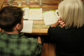 A man in a green plaid shirt and a woman with short blonde hair sit at a wooden table, looking at open books or study guides in a warm, indoor setting.