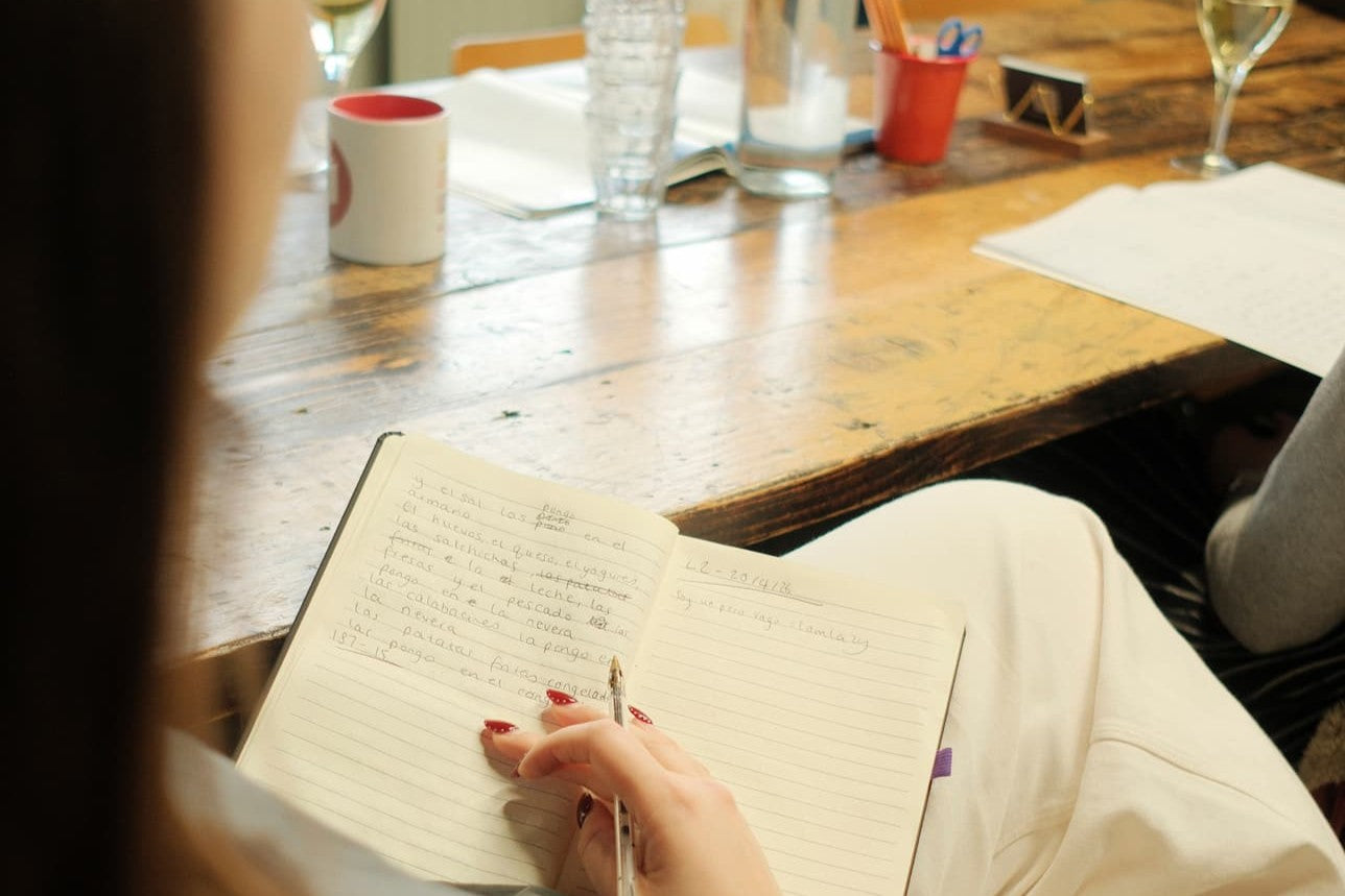A person writing in a lined notebook with a pen, seated at a rustic wooden table with mugs, glasses, and papers in a cafe or meeting setting.