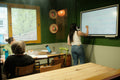 A woman stands at a whiteboard in a classroom or meeting space, writing on the board while other people sit at tables in the foreground. The room has dark green walls, a large window, and a wooden table in the immediate foreground.