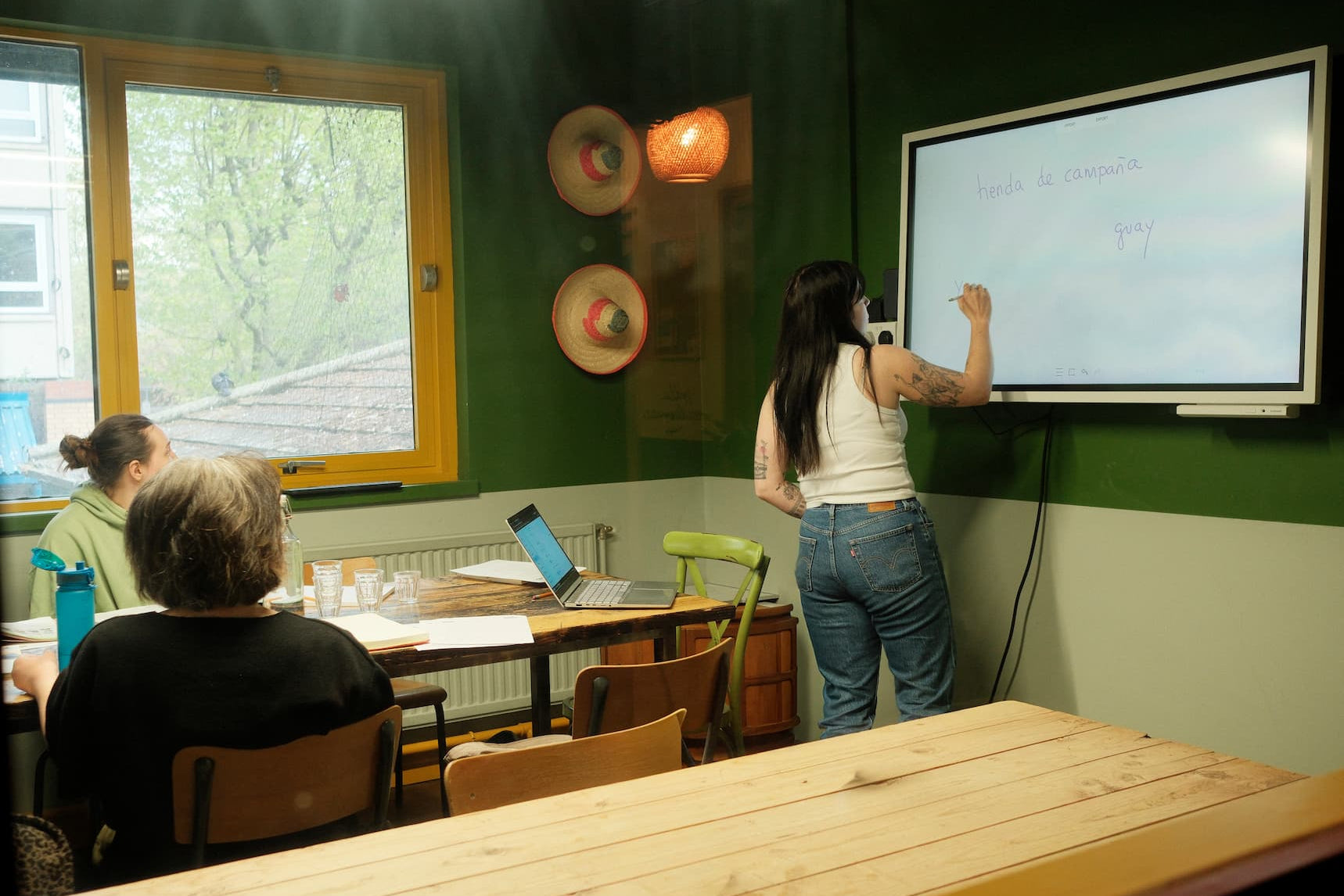 A woman stands at a whiteboard in a classroom or meeting space, writing on the board while other people sit at tables in the foreground. The room has dark green walls, a large window, and a wooden table in the immediate foreground.