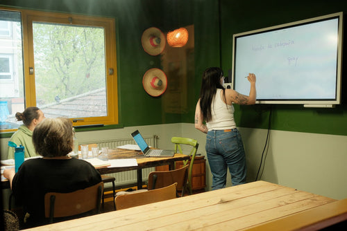 A woman stands at a whiteboard in a classroom or meeting space, writing on the board while other people sit at tables in the foreground. The room has dark green walls, a large window, and a wooden table in the immediate foreground.