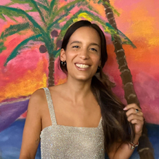 A smiling woman with long dark hair stands in front of a colorful painted tropical sunset scene, wearing a sparkly silver sleeveless top and blue jeans.