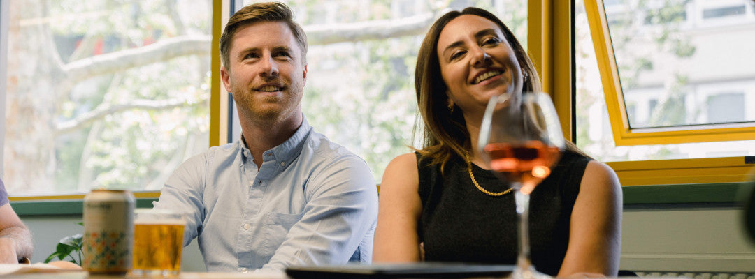 A smiling man in a light blue button-down shirt and a woman in a black sleeveless top sitting at a table by a window, with a glass of rosé wine in the foreground.