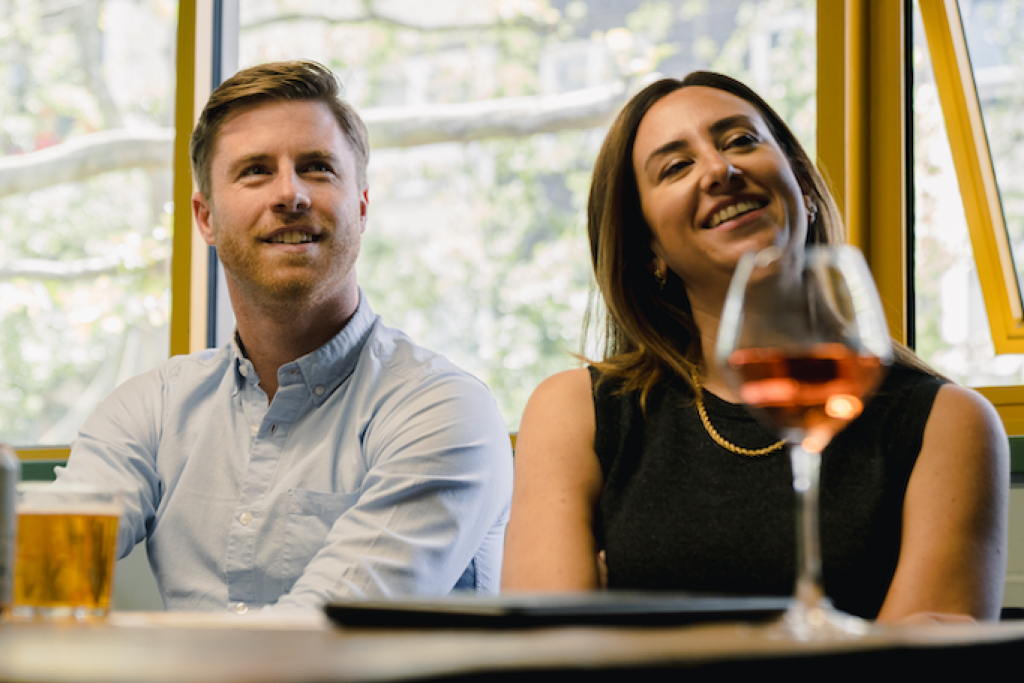 A man and a woman sitting at a table in a bright indoor setting, smiling and looking toward the camera. A glass of pink wine and a beer glass are in the foreground, with large windows and trees visible behind them.