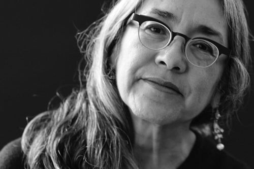 A black and white close-up portrait of a woman with long hair and glasses, wearing dangling earrings against a dark background.