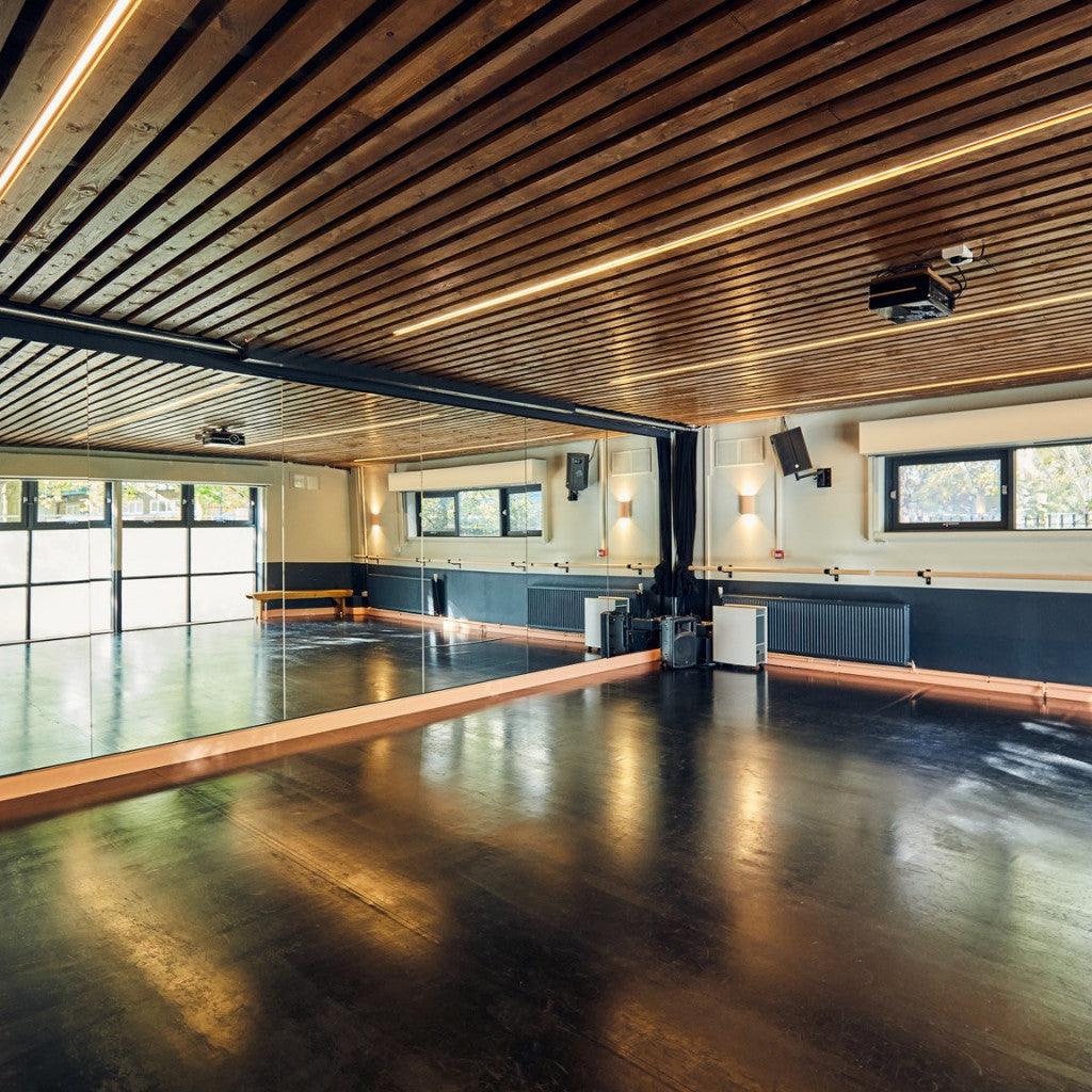 An empty modern dance studio with a dark wood slatted ceiling, warm LED strip lighting, a glossy dark floor, mirrored walls, windows, and wall bars.