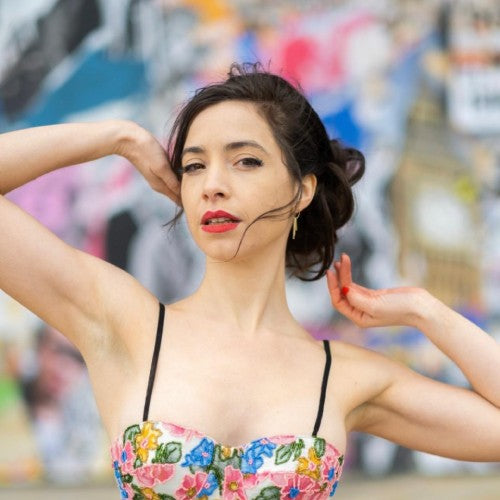 A woman with dark hair in an updo wearing a colorful floral strapless top with thin black straps, posing with her arms raised against a blurred graffiti background.