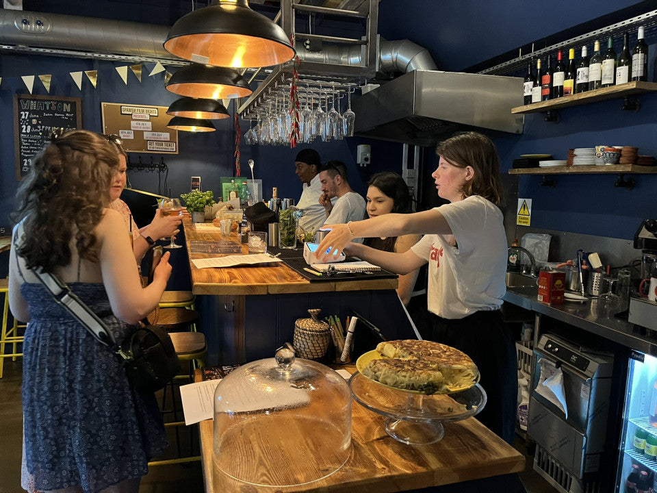 A woman in a white t-shirt and black skirt points toward a menu or board at a wooden bar inside a dark blue restaurant. Another woman in a blue patterned dress stands on the left, and other staff and patrons are visible in the background with pendant lights, wine bottles, and a food display in the foreground.
