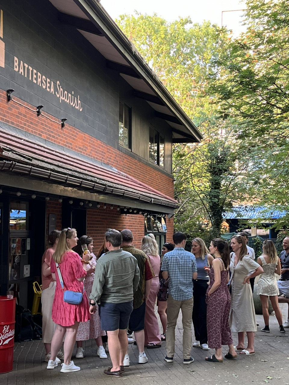 A group of people standing in a queue outside a restaurant or bar with a dark facade and brick lower section. Trees and daylight are visible in the background, and signage on the building reads 'BATTERSEA Spanish'.