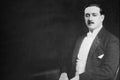 Black and white portrait photograph of a man with a mustache, wearing a dark suit jacket and white shirt against a dark studio background.