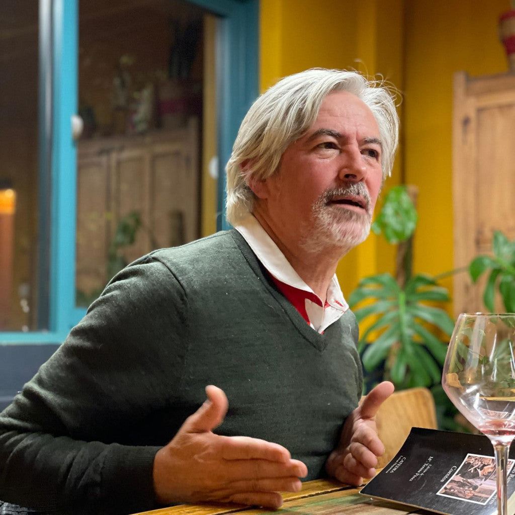 A man with gray hair and a mustache wearing a dark green V-neck sweater over a white and red collared shirt, sitting at a table with a wine glass and a menu in a warm, indoor setting.