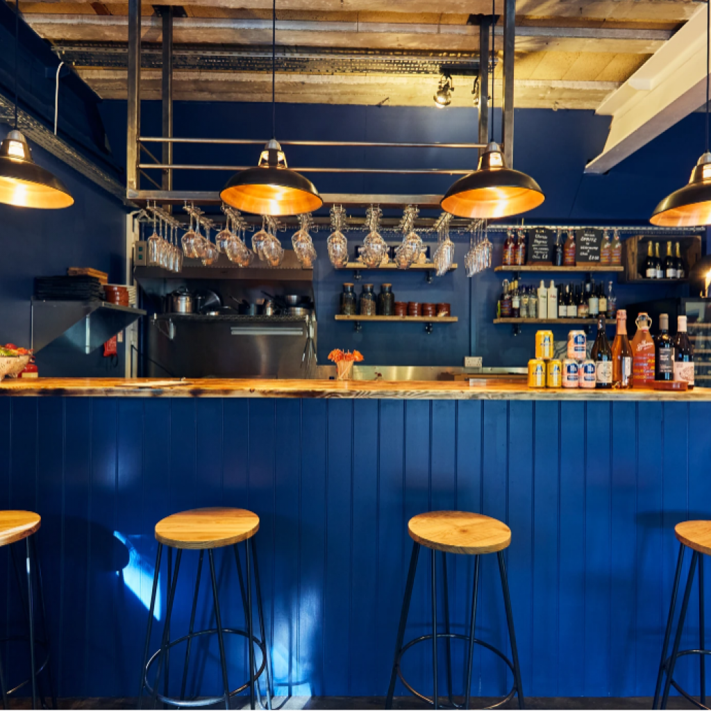 A blue bar interior with a wood counter, hanging bar stools, pendant lights, and shelves stocked with bottles and glassware.