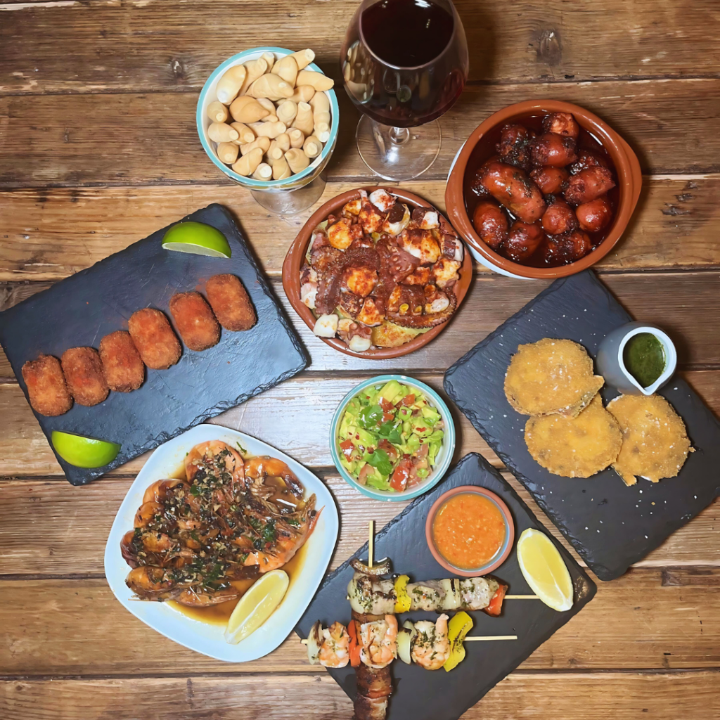 A wooden table styled with a full Mexican meal spread, including tacos, guacamole, salsa, chorizo, mole, peanuts, and various skewers, arranged in a flat-lay style.