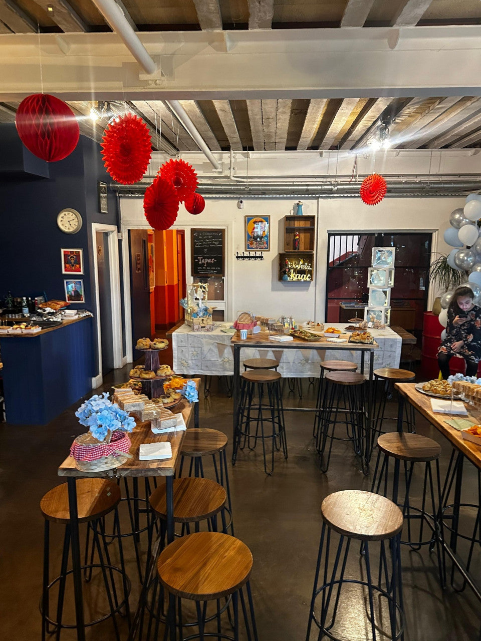 A decorated indoor event space or party venue with multiple wooden bar stools, tables, hanging red paper lanterns, and a reception-style food table in the background.