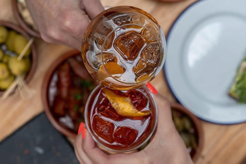 Two hands holding up two clear glasses of iced red cocktail with ice cubes and orange garnish, over a wooden table with food bowls and a white plate.