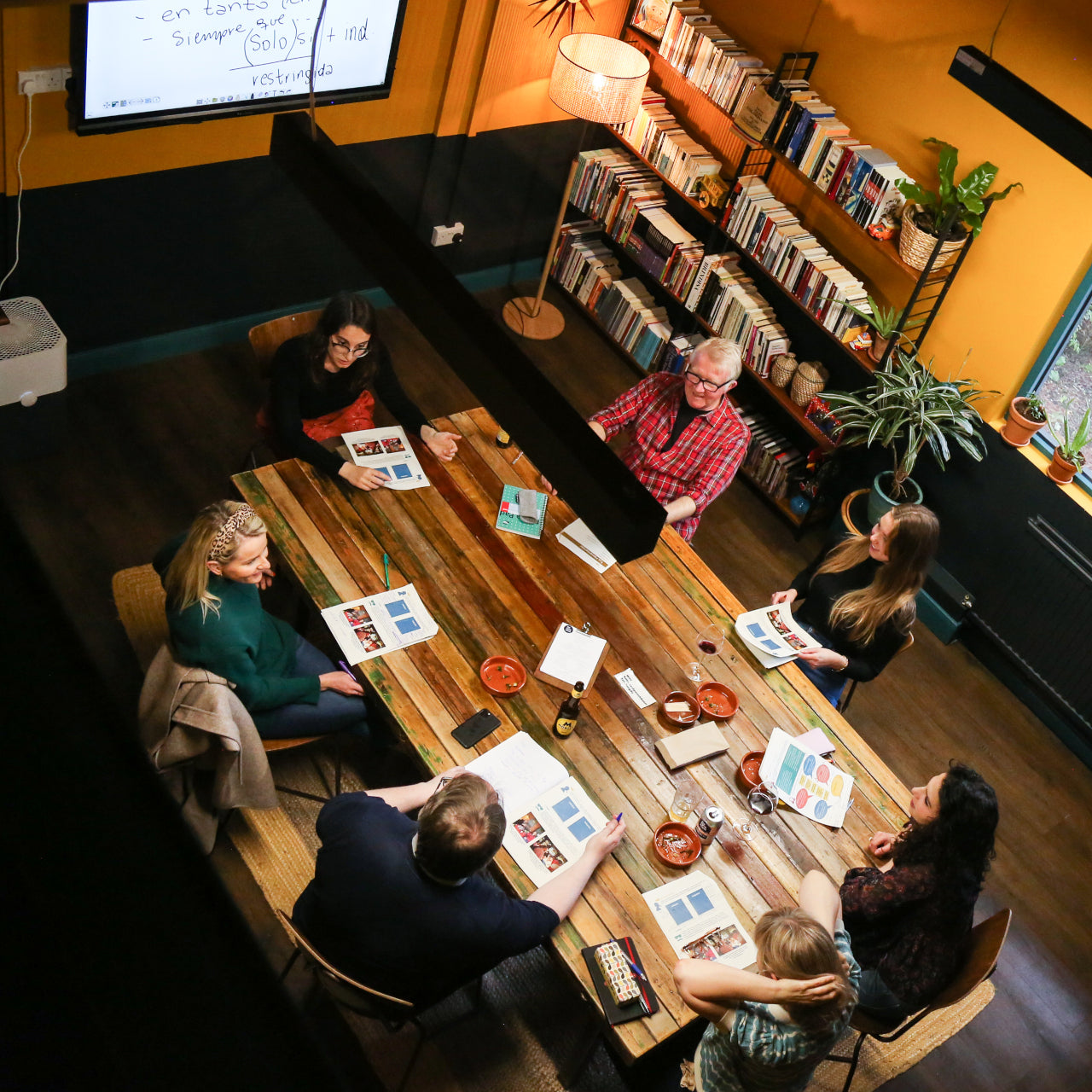 An overhead view of a group of people seated around a wooden table in a warm, book-filled room, looking at printed materials and engaging in discussion. Bookshelves, plants, a floor lamp, and a projector screen are visible in the background.