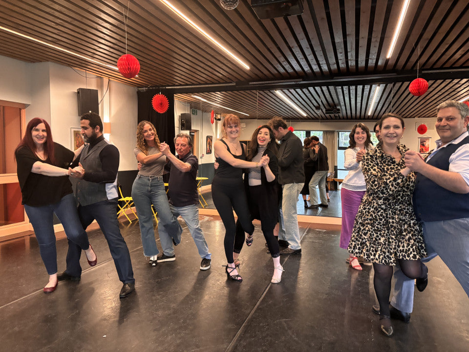A group of people dancing together in a dance studio with wooden ceiling panels, red paper lanterns hanging from the ceiling, and a dark floor.